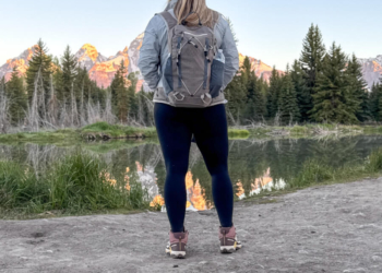 Woman stands facing away from camera, overlooking a lake, pine trees and mountains.