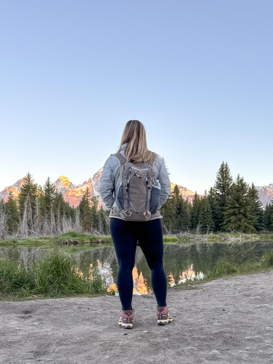 Woman stands facing away from camera, overlooking a lake, pine trees and mountains.