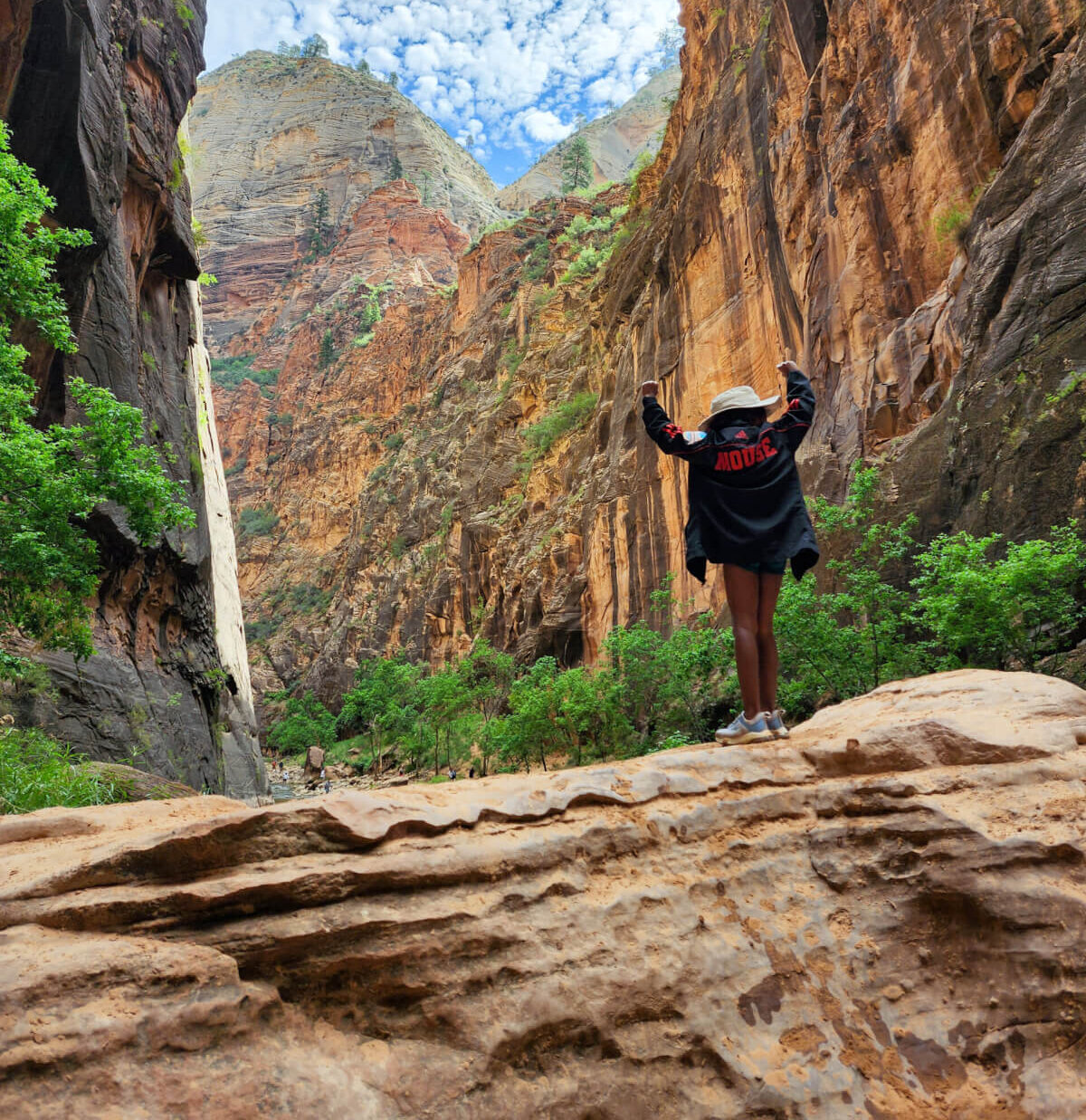 Military kid and taekwondo athlete Madison Richardson stands in a canyon on a rock, facing away from the camera, with her arms held high in victory. The back of her jacket reads "Mouse," in reference to her nickname, "MightyMouse."