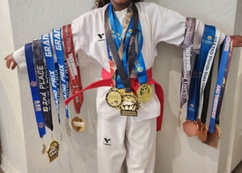 Military kid and taekwondo athlete Madison Richardson stands in her white dobok uniform, with her arms outstretched. She has medals around her neck and each of her arms, and is smiling broadly.