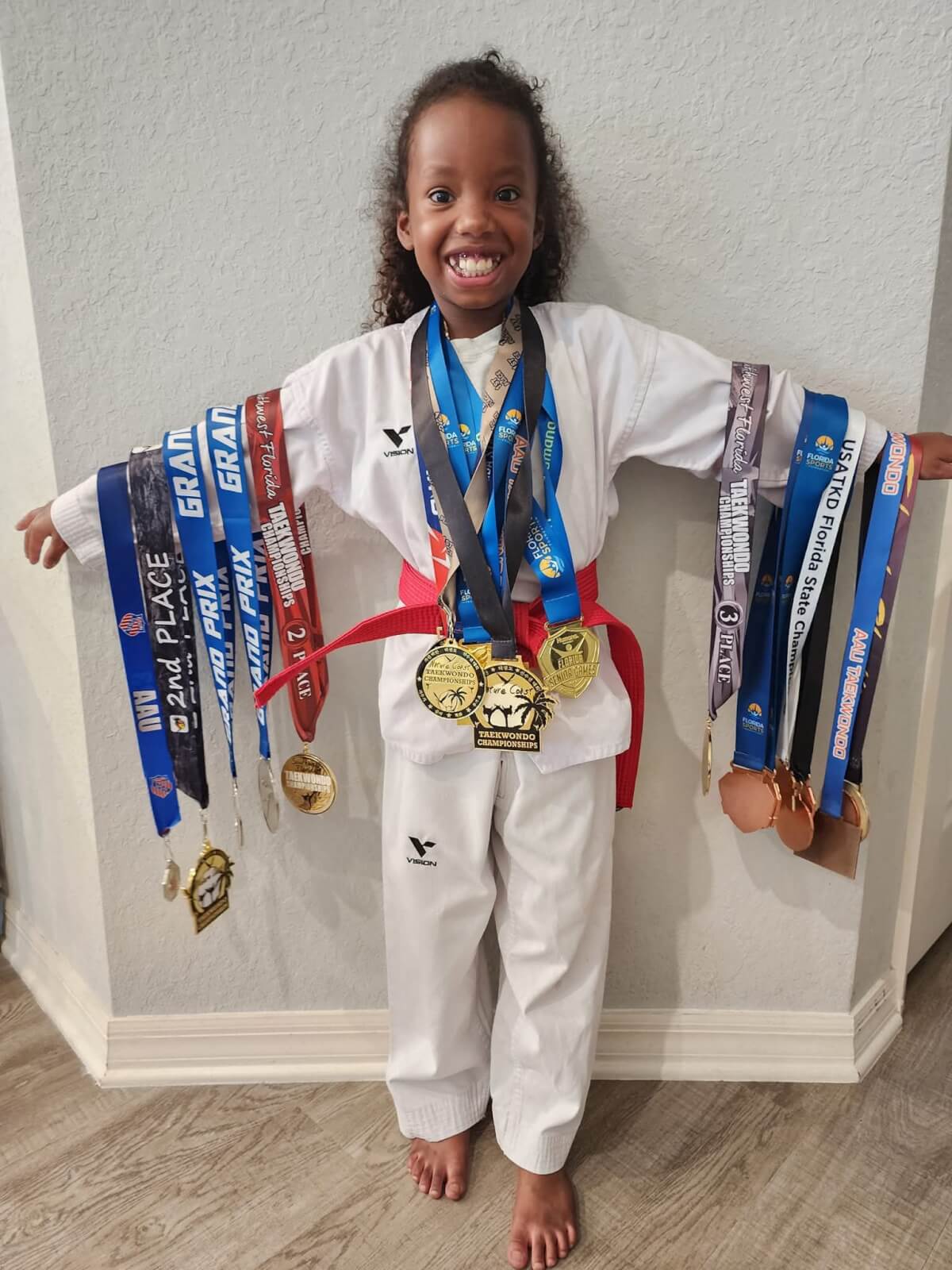 Military kid and taekwondo athlete Madison Richardson stands in her white dobok uniform, with her arms outstretched. She has medals around her neck and each of her arms, and is smiling broadly.
