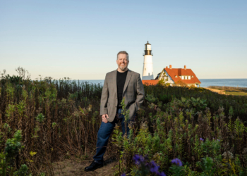 Boots2Roots Executive Director Randy Bell stands among tall grass with a charming lighthouse and water in the background.