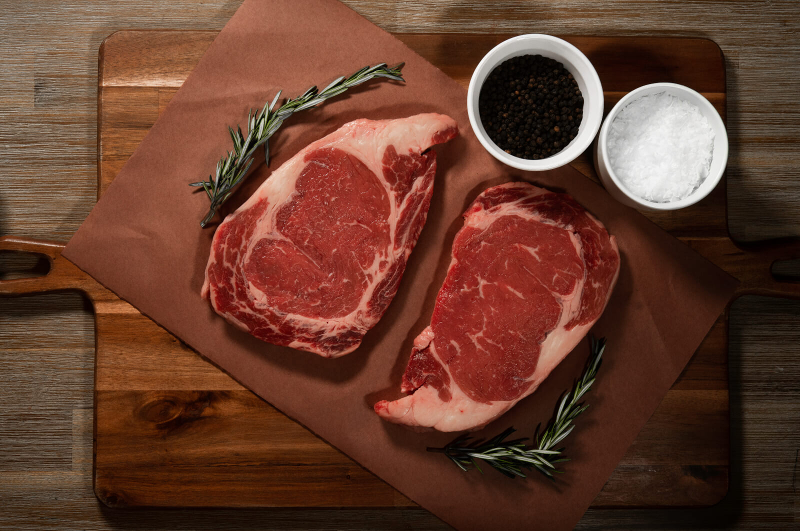 Two red steaks, from 8Primal, a military-owned small business, on a cutting board with herbs, salt and pepper around them.