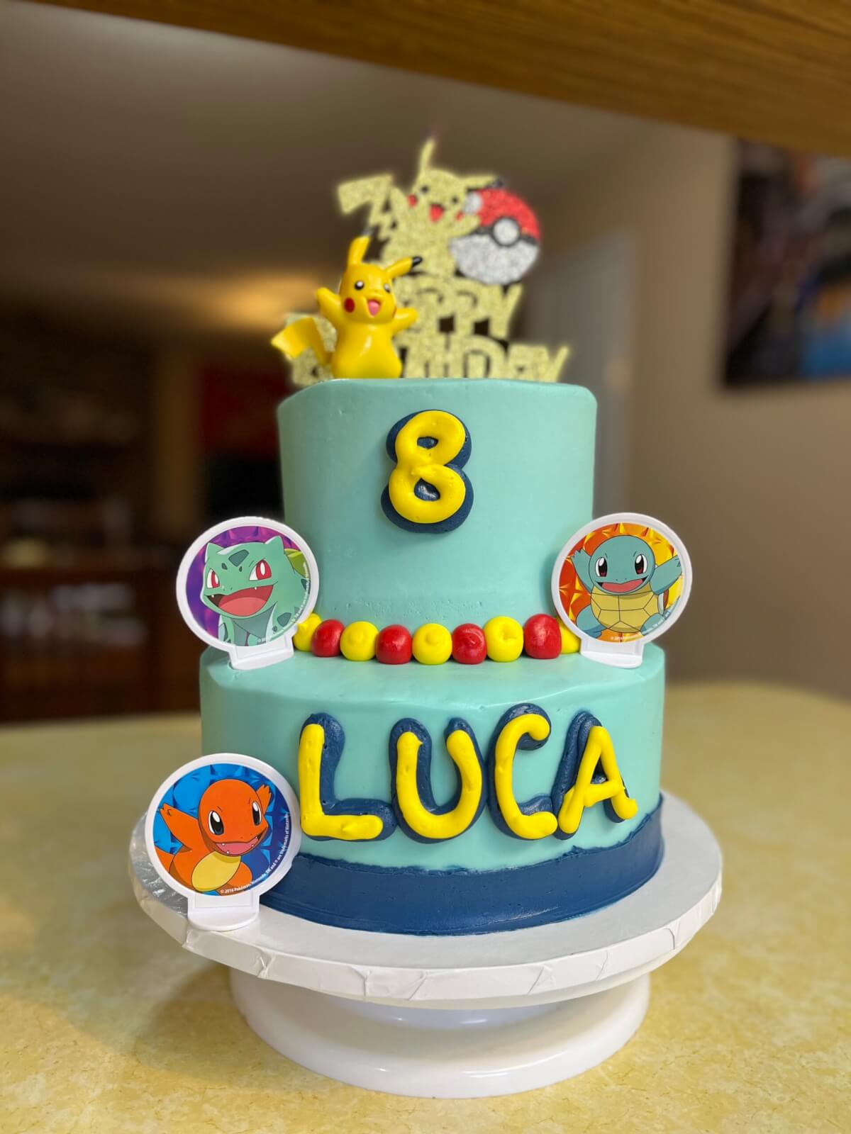 Two-tiered birthday cake made by Marine spouse baker. Cake is blue with yellow and red decorations, and yellow piping spells out 'LUCA.' Pokemon characters are on each tier.