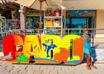 Military child smiling in his sunglasses as he leans against a brightly colored "CABO" sign on vacation in Mexico.