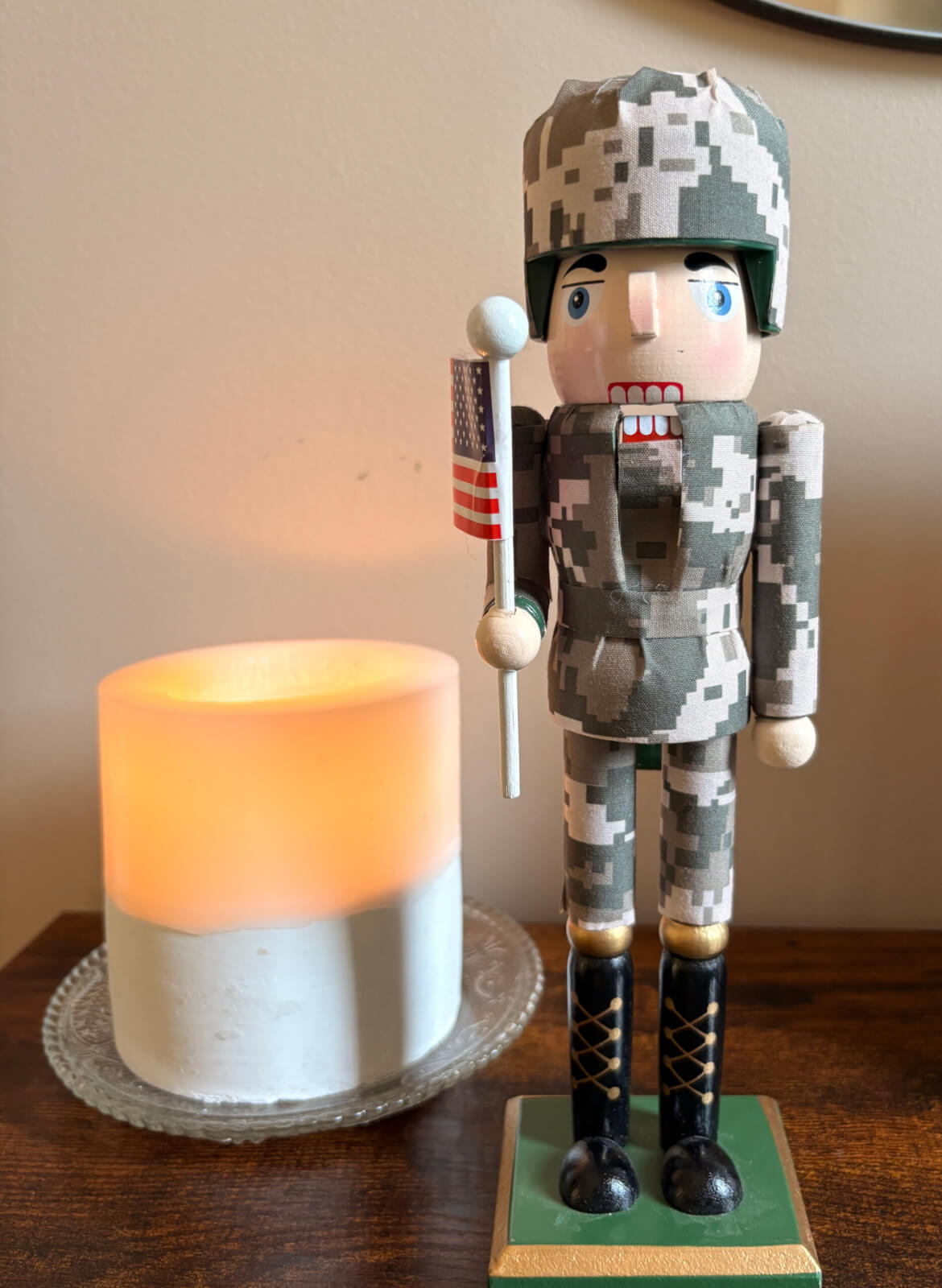 Next to a cozy lit candle on a table, a wooden nutcracker in a military uniform holds an American flag as patriotic holiday décor.