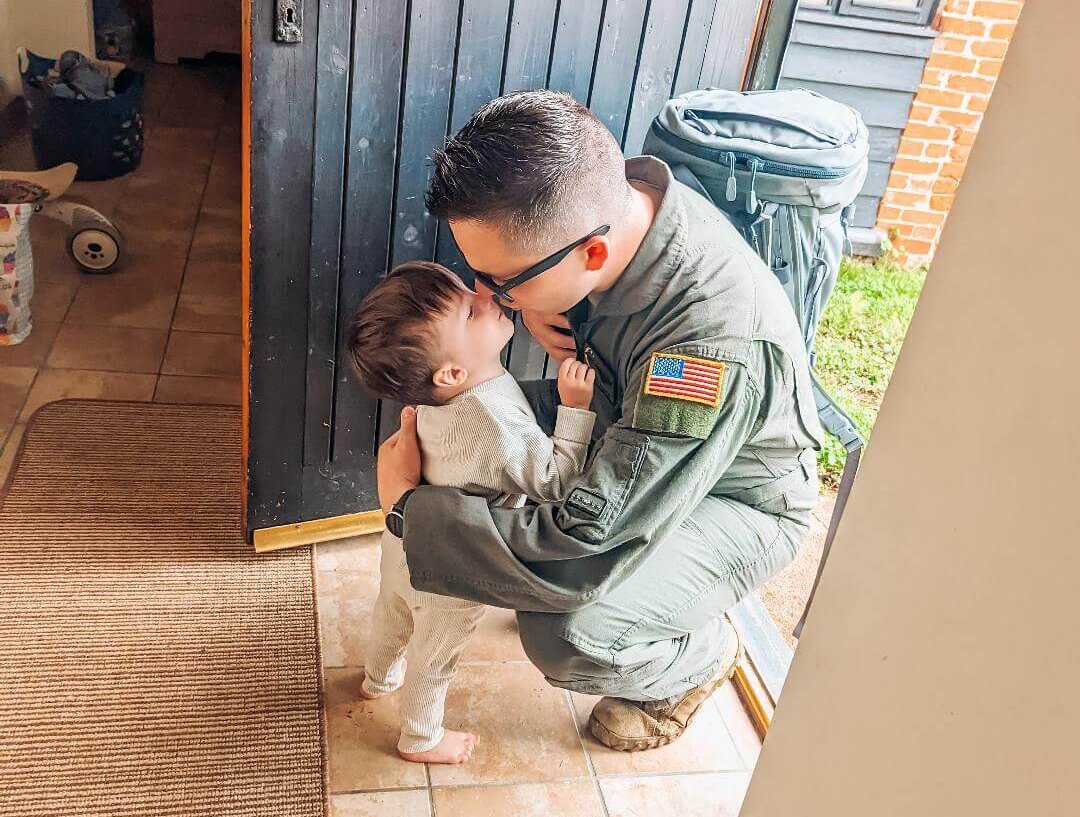 Air Force father in uniform squats to kiss young toddler son goodbye as he leaves the house.