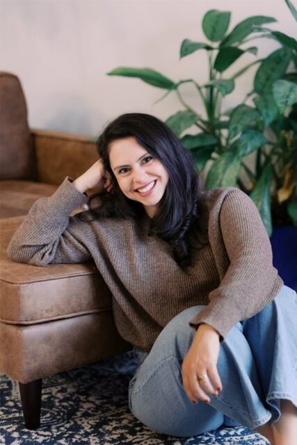 With a green plant in the background, poet Hannah Rosenberg sits on a carpeted floor, leaning with her back and elbow against a brown leather chair. 
