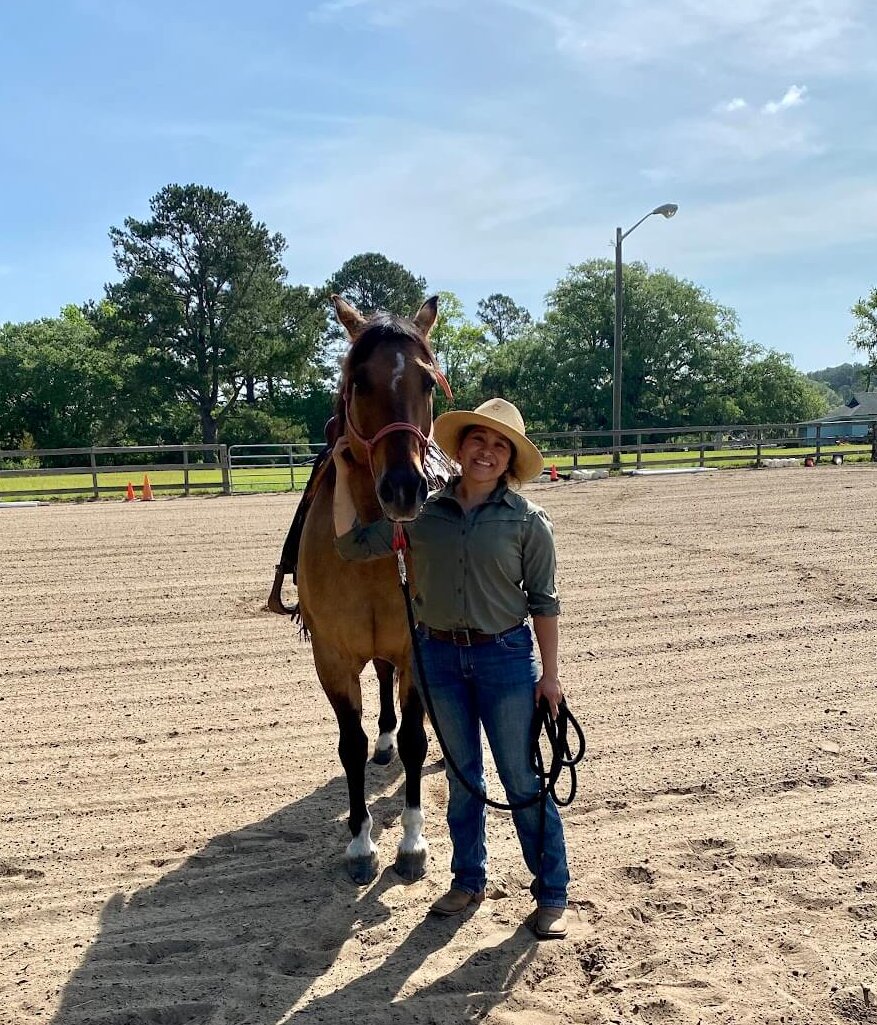 As a Tillman Foundation Scholar, Nina Guerra supports veterans with equine therapy. Wearing a green shirt, jeans, and boots, she holds the rope of the brown horse beside her. 
