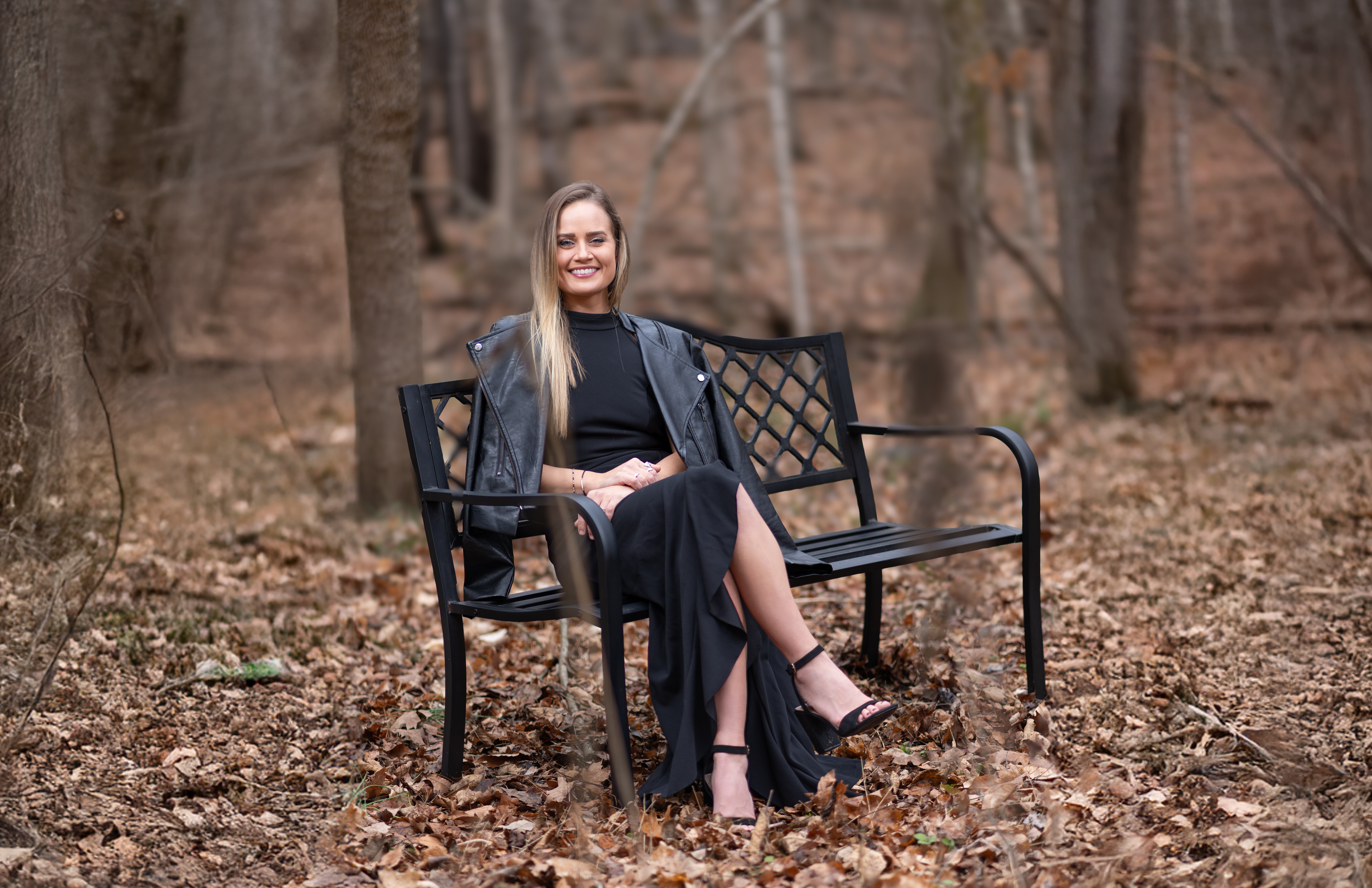 Ashlee Sullivan of WOMAN + WARRIOR sits outside on a black bench surrounded by fallen leaves.
