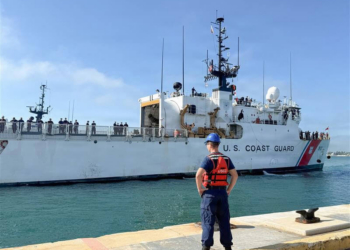 With backdrop of blue sky and on blue-green water, Coast Guard Cutter Mohawk approaches the pier at its homeport in Key West, Florida, as a Coast Guardsman stands on the pier and looks on.