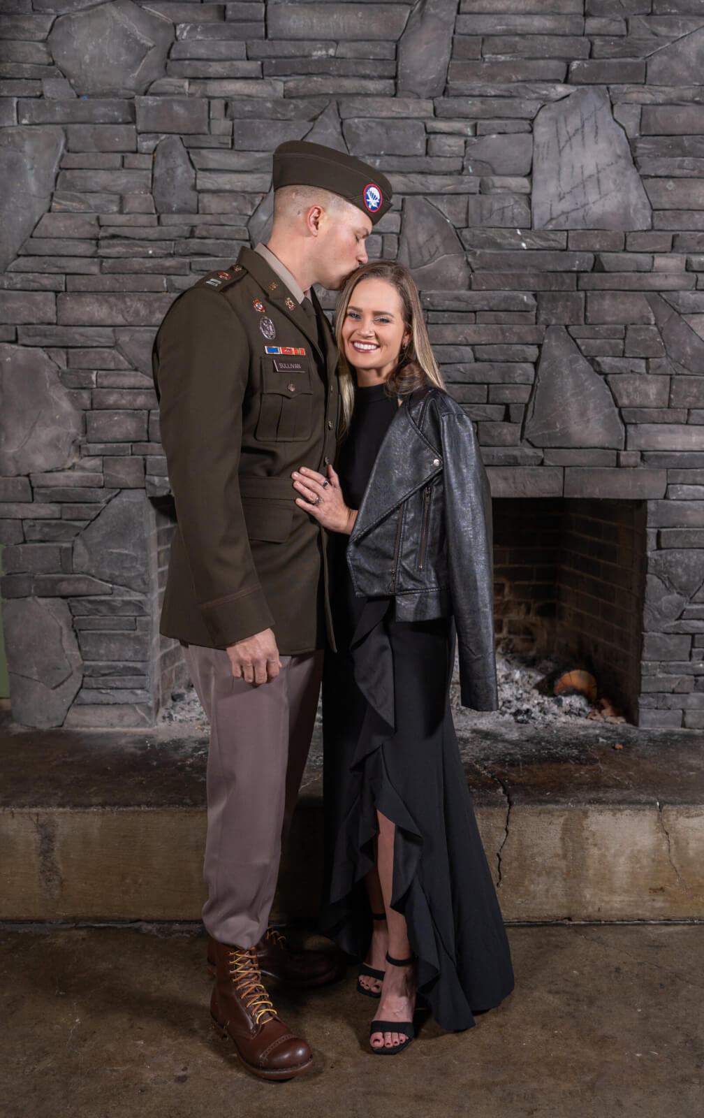 Wearing his Army uniform, Capt. Peter Sullivan kisses his wife Ashlee of WOMAN + WARRIOR in front of a stone fireplace.