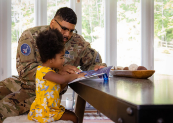 United Through Reading encourages parents and caregivers to read to children at home. A Space Force father in uniform reads to his daughter at the kitchen table.