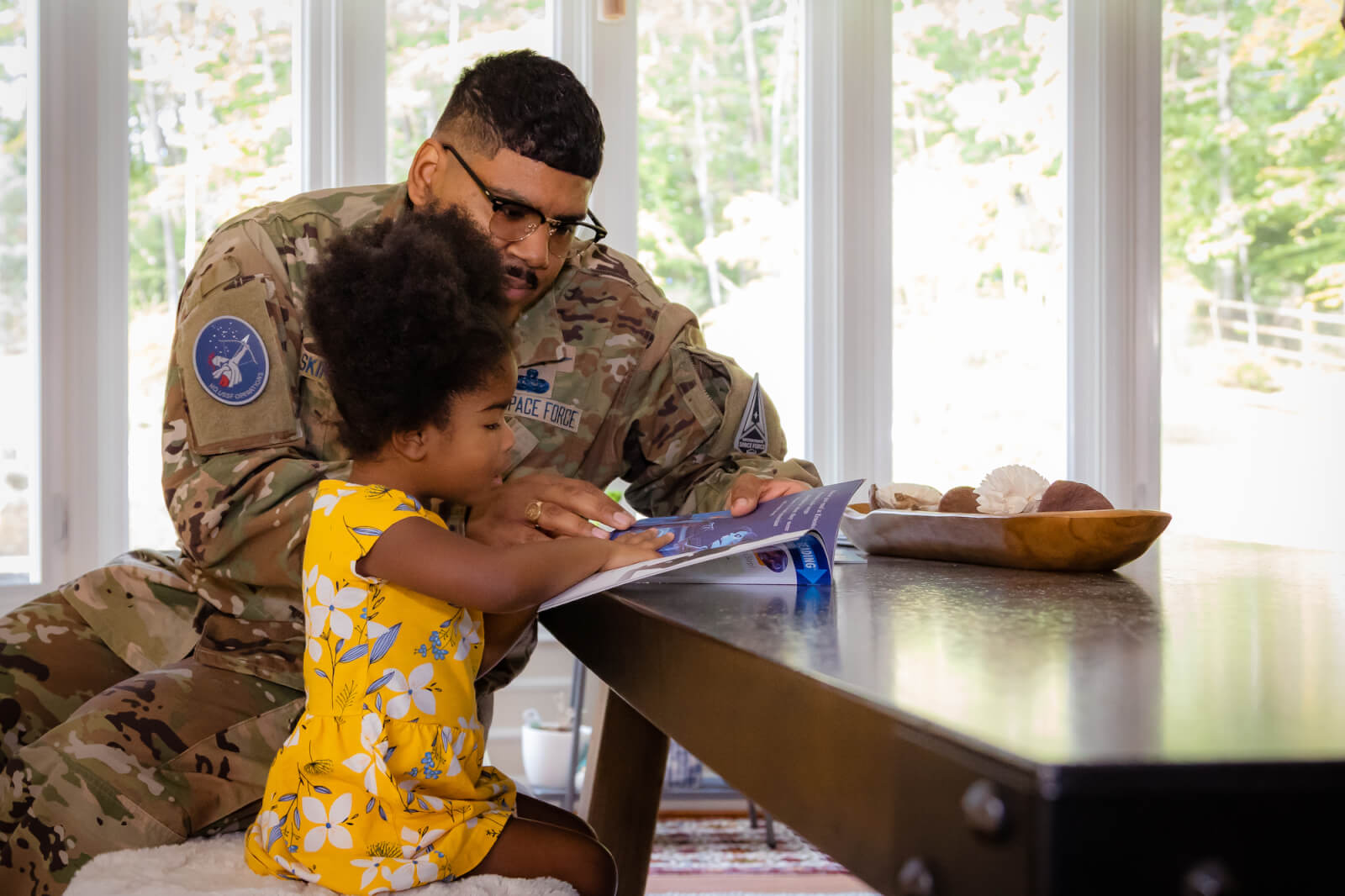 United Through Reading encourages parents and caregivers to read to children at home. A Space Force father in uniform reads to his daughter at the kitchen table.