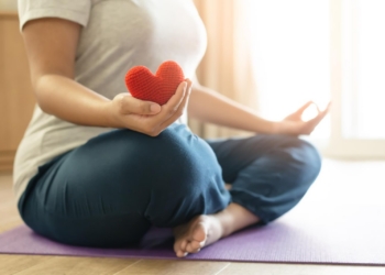 Woman practices self care with meditation and yoga, holding a red yarn heart.
