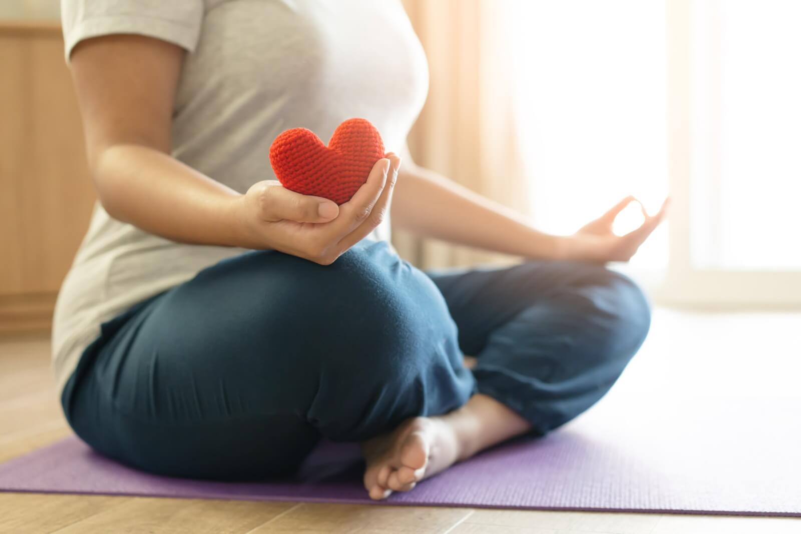Woman practices self care with meditation and yoga, holding a red yarn heart.