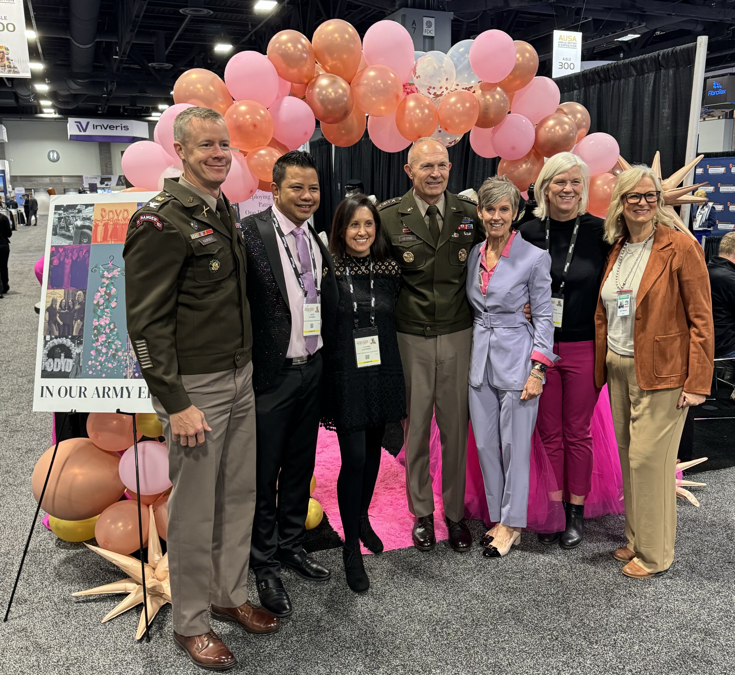 Seven people —two military members in uniform— stand in front of an Operation Deploy Your Dress booth with a pink and coral balloon arch behind them.