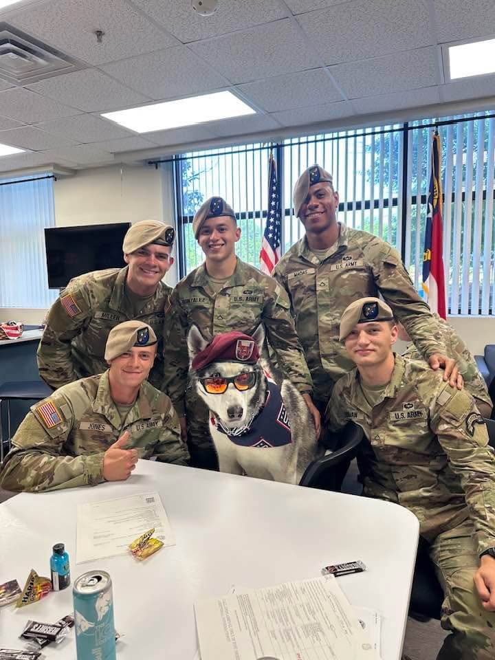 Sapper sits in the middle of five service members in uniform. Sapper wears his sunglasses, maroon beret, and USO bandana.