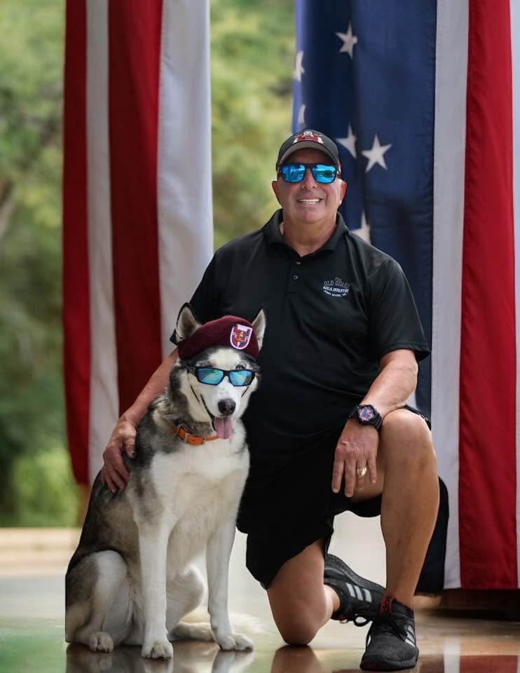 Sapper sits next to his handler Mike. Both are wearing sunglasses, and Sapper is wearing a maroon beret.