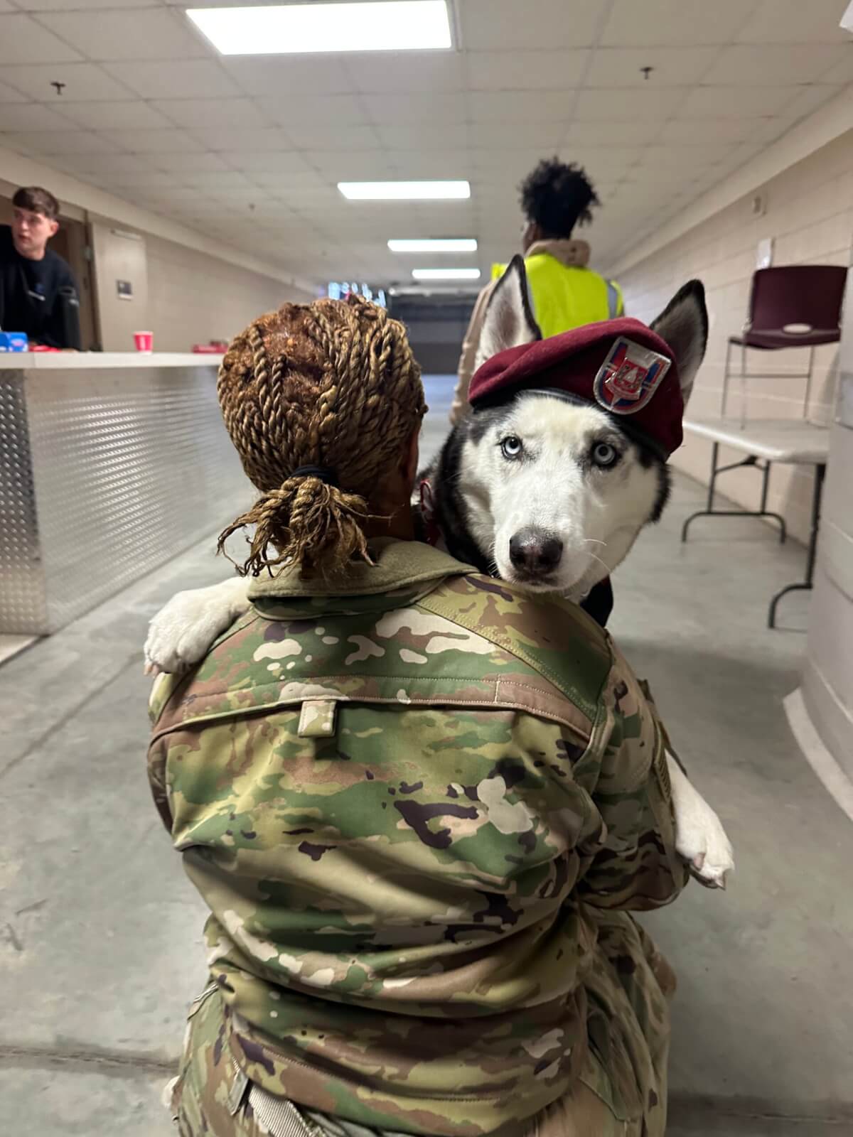 A female service members hugs Sapper, with him looking over her shoulder. He is wearing his maroon military beret.