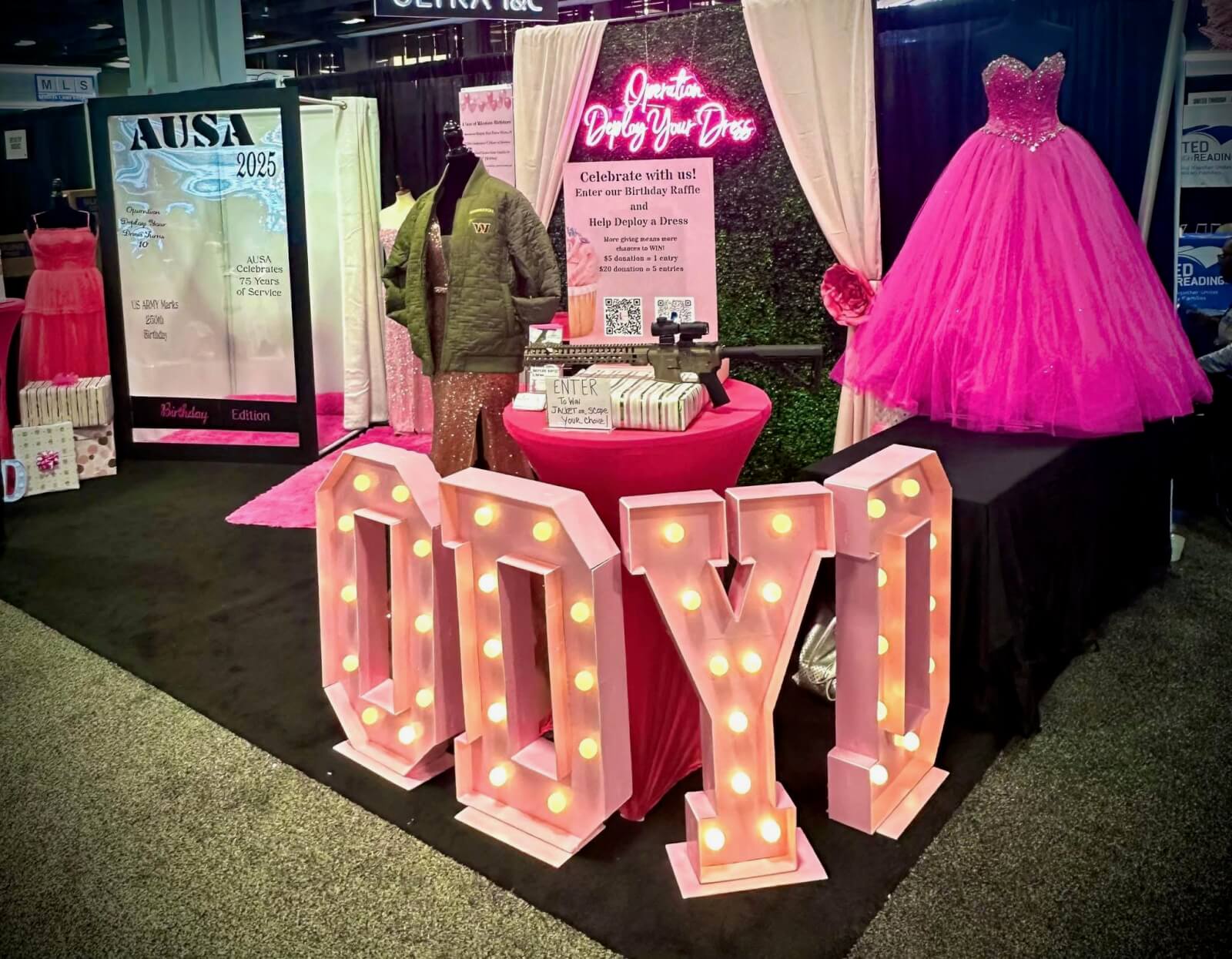 Operation Deploy Your Dress booth with large pink ODYD letters lit up with lights, with pink curtains and a pink dress in the background.