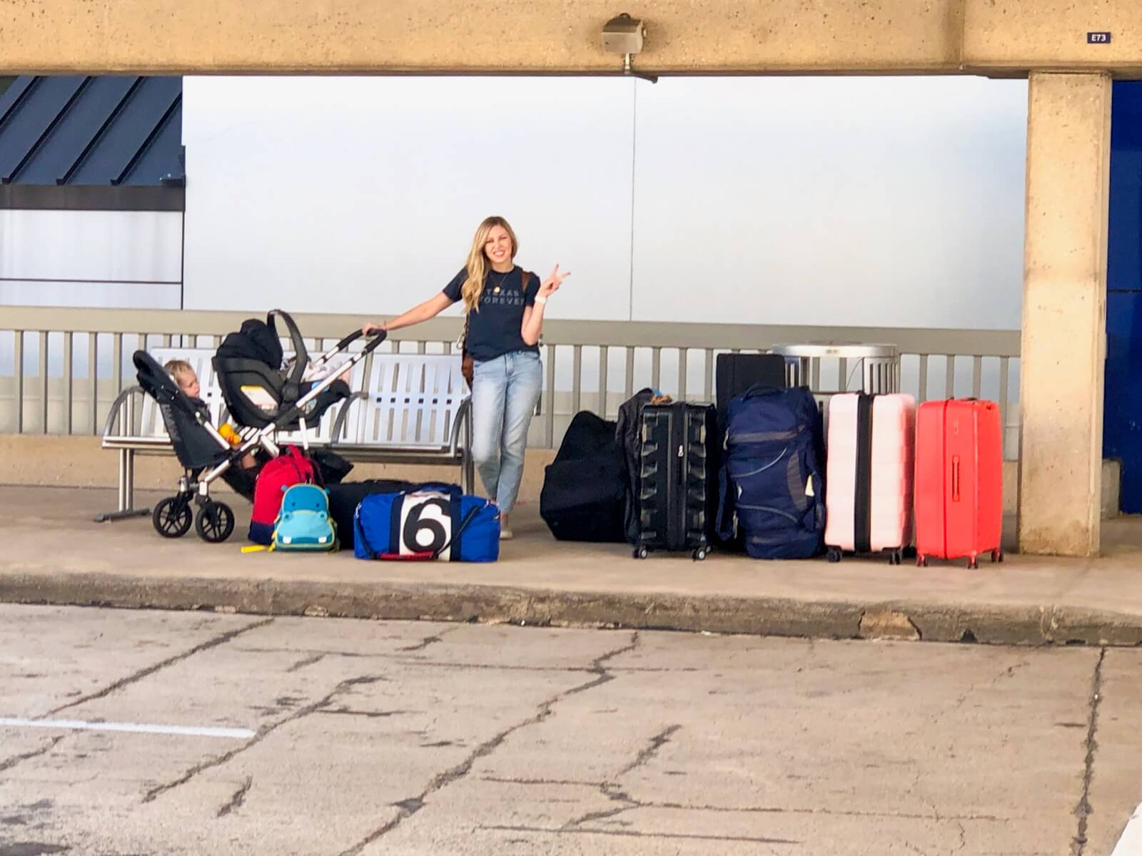 Woman stands with piles of luggage and a stroller with a baby while making a peace sign.